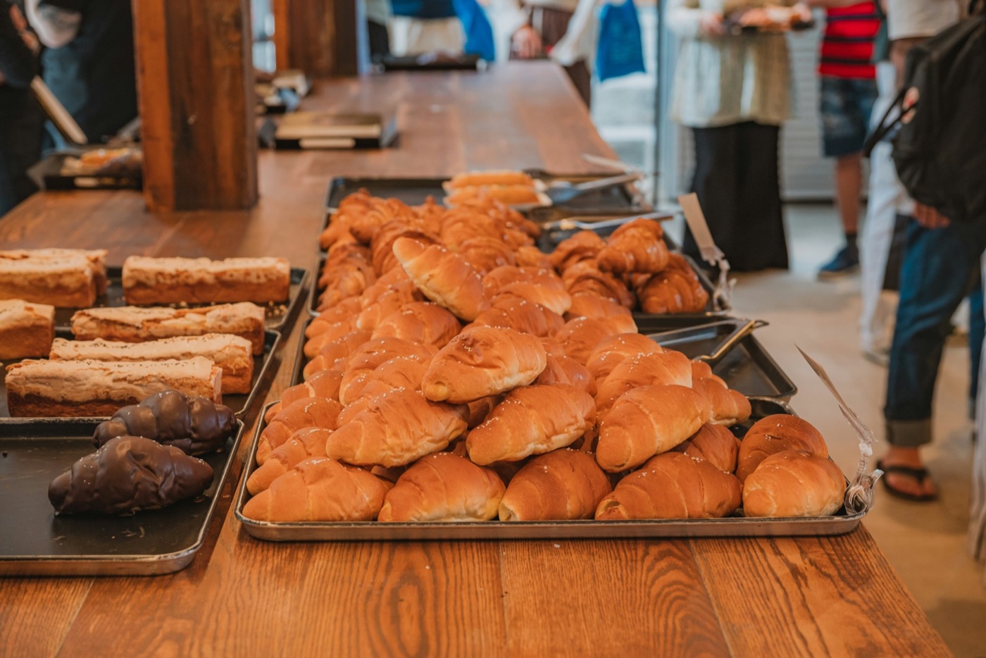 Trays of fresh rolls, loaf cakes and chocolate pastries on a long wooden bakery counter, with customers in the background