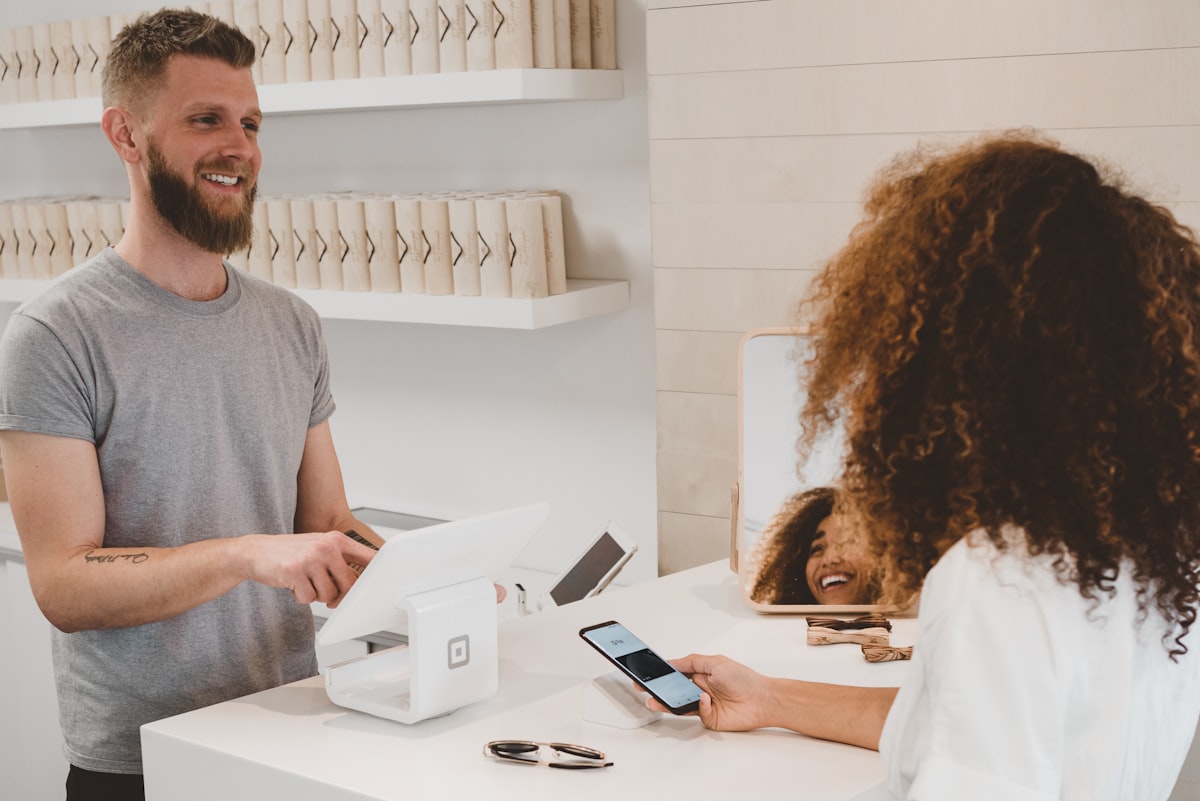 A customer holds a phone at a shop counter while the owner behind a card terminal smiles